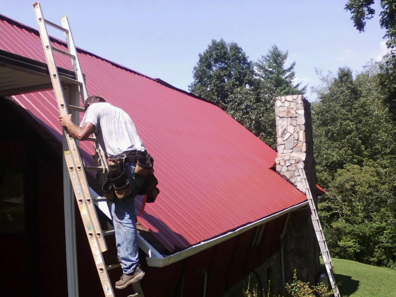 Roofer on a ladder beside a red metal roof and stone chimney — Soffit & Fascia Repair by Roofing Brothers in Fort George G Meade